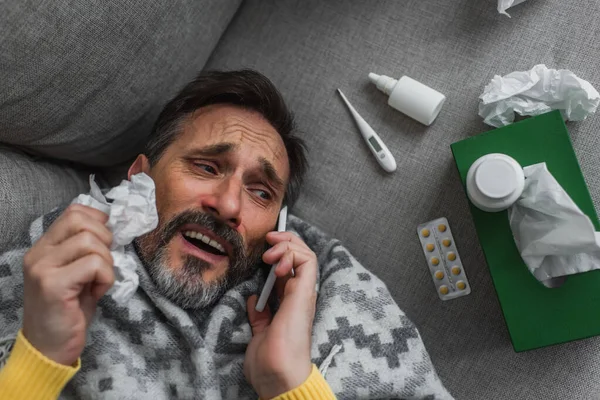 Vue du dessus de l'homme malade parlant sur smartphone tout en étant couché près du thermomètre et des pilules sur le canapé — Photo de stock