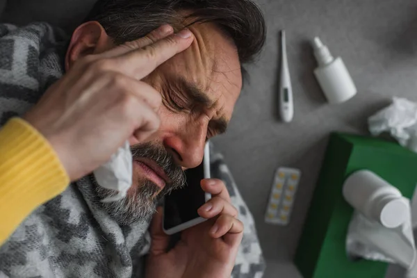 Vista superior del hombre enfermo que sufre de fiebre y dolor de cabeza mientras llama a un teléfono inteligente cerca de pastillas y contenedores con tratamiento - foto de stock