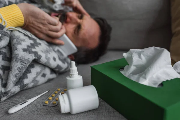 Electronic thermometer and containers with treatment near sick man lying on couch and calling on smartphone — Stock Photo