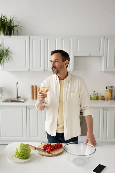 Uomo barbuto in camicia bianca in possesso di un bicchiere di vino e guardando lontano vicino a pomodori ciliegini maturi e lattuga fresca — Foto stock