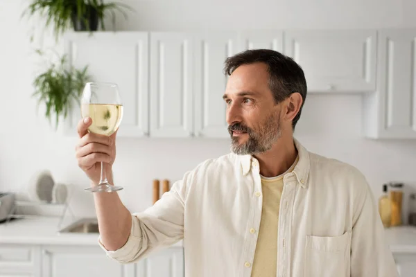 Uomo sorridente con barba grigia che guarda un bicchiere di vino bianco in cucina — Foto stock