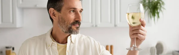 Bearded man in white shirt looking at glass of white wine in kitchen, banner — Stock Photo