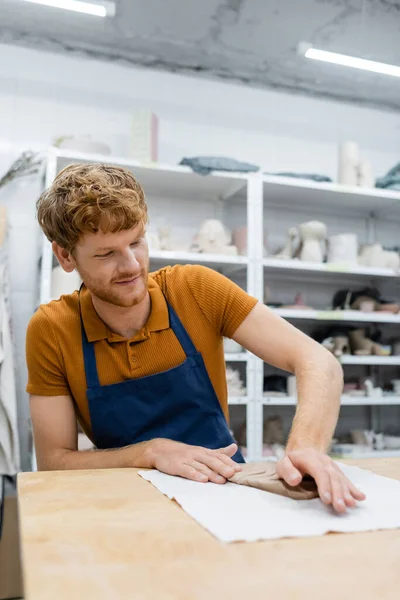 Hombre pelirrojo feliz en delantal presionando la pieza de arcilla con la mano durante la clase de cerámica — Stock Photo