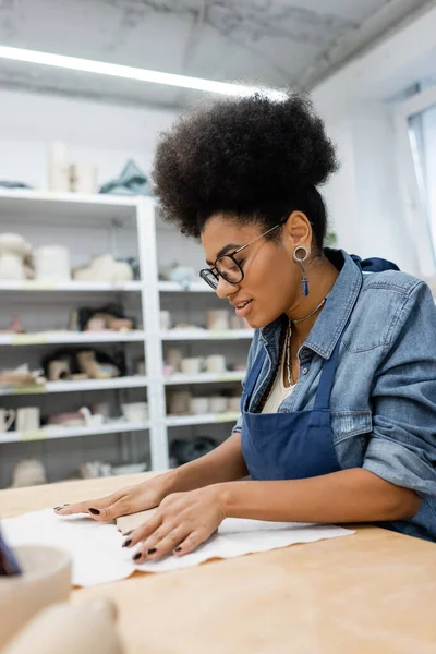 Кудрявая африканская американка в фартуке и очках, с руками в керамической мастерской — стоковое фото