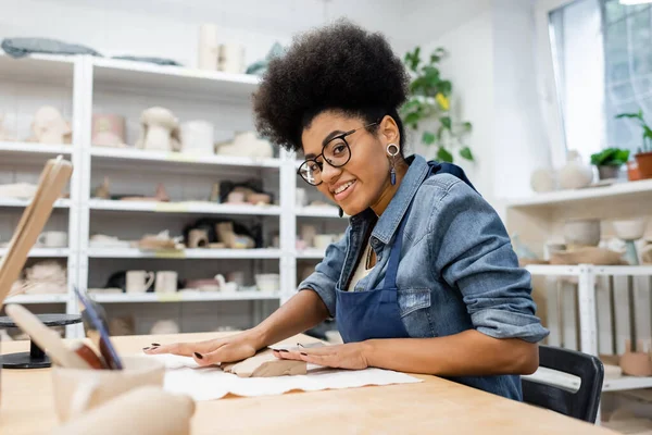 Femme afro-américaine gaie dans tablier et lunettes façonner morceau d'argile avec les mains pendant le cours de poterie — Photo de stock