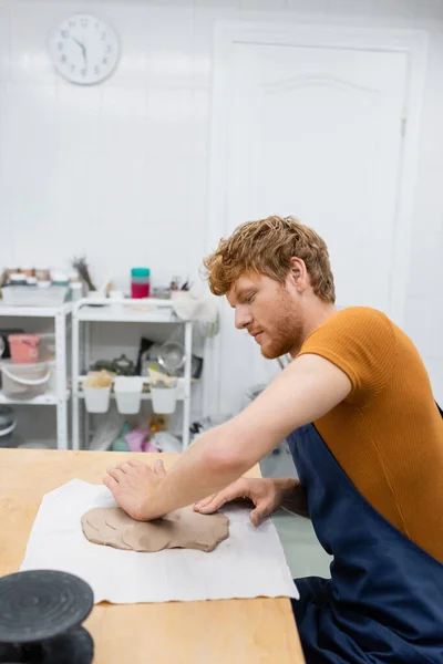 Vista lateral do homem ruivo no avental moldando peça de argila com a mão em oficina de cerâmica — Fotografia de Stock