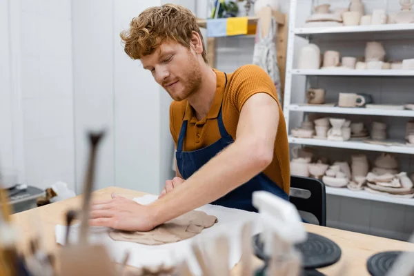 Bearded redhead man in apron shaping clay piece with hand in pottery workshop — Stock Photo