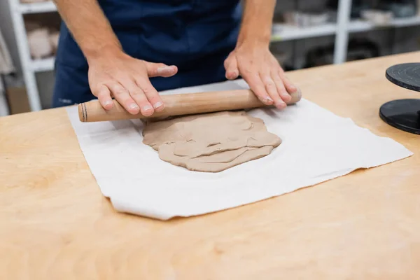 Cropped view of man in apron shaping clay piece with rolling pin during pottery class — Stock Photo