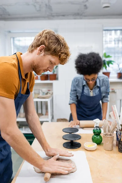 Redhead man in apron shaping clay piece with rolling pin near african american woman during pottery class — Stock Photo