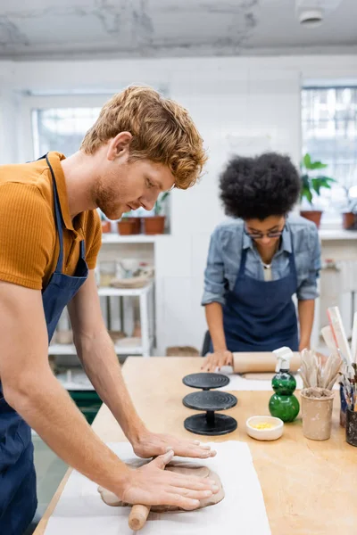 Pelirrojo en delantal dando forma a la pieza de arcilla con rodillo cerca de la mujer afroamericana durante la clase de cerámica — Stock Photo