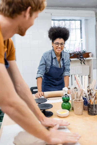 Heureuse femme afro-américaine en lunettes façonnant morceau d'argile avec rouleau à pâtisserie près de rousse homme sur le premier plan flou — Photo de stock