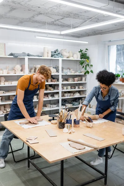 Interracial couple in aprons shaping clay with rolling pins in pottery workshop — Stock Photo
