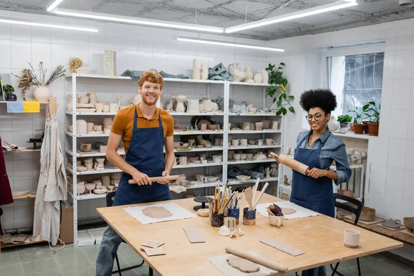 Glückliches gemischtes Paar in Schürzen mit Nudelholz in Töpferwerkstatt — Stockfoto