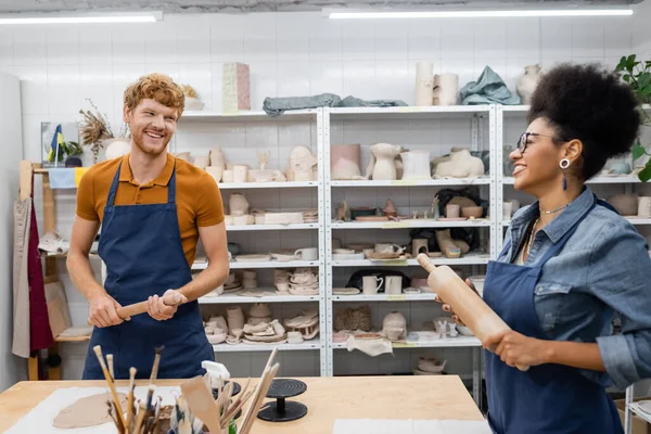 Happy interracial couple in aprons holding rolling pins while looking at each other during pottery class — Stock Photo