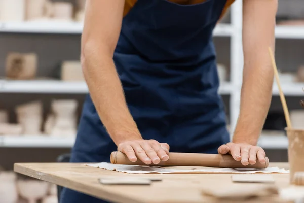 Vista recortada del hombre en delantal modelado pieza de arcilla con rodillo en taller de cerámica - foto de stock