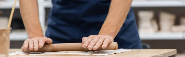 Vue recadrée de l'homme en tablier pièce d'argile de modélisation avec rouleau à pâtisserie pendant la classe de poterie, bannière — Photo de stock