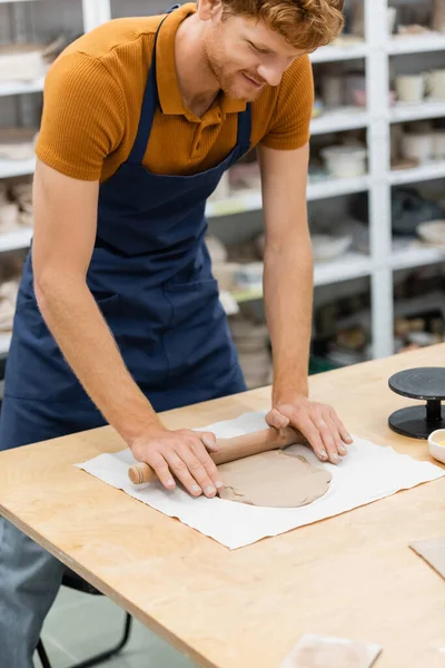 Alegre pelirrojo hombre en delantal modelado pieza de arcilla con rodillo durante la clase de cerámica — Stock Photo