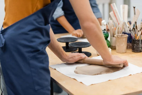 Cropped view of man in apron modeling clay piece with rolling pin near woman during pottery class — Stock Photo