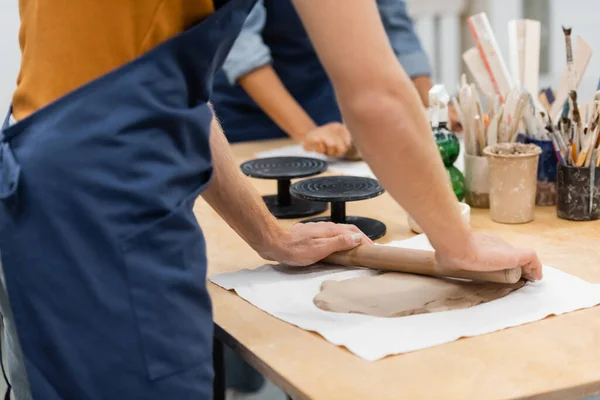 Vista recortada del hombre en delantal modelado pieza de arcilla con rodillo cerca de la mujer durante la clase de cerámica — Stock Photo