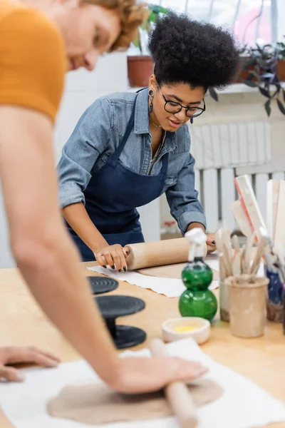 African american woman in eyeglasses and apron modeling clay with rolling pin near man — Stock Photo