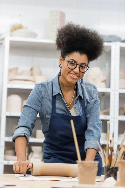 Cheerful african american woman in eyeglasses and apron modeling clay with rolling pin — Stock Photo