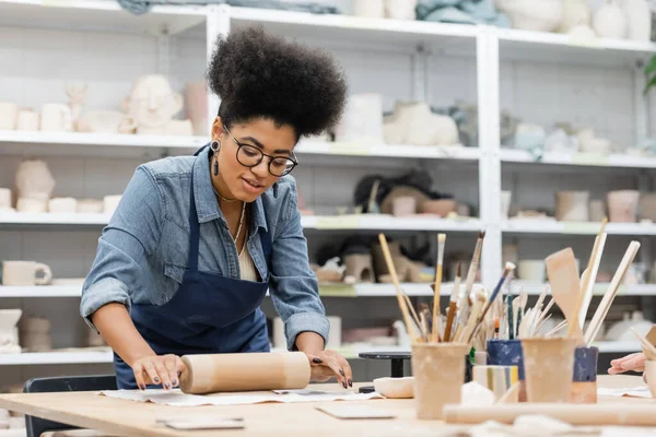 Joven afroamericana mujer en gafas y delantal modelado de arcilla con rodillo — Stock Photo