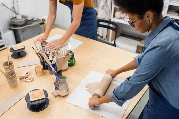 High angle view of african american woman in eyeglasses modeling clay piece with rolling pin near man — Stock Photo