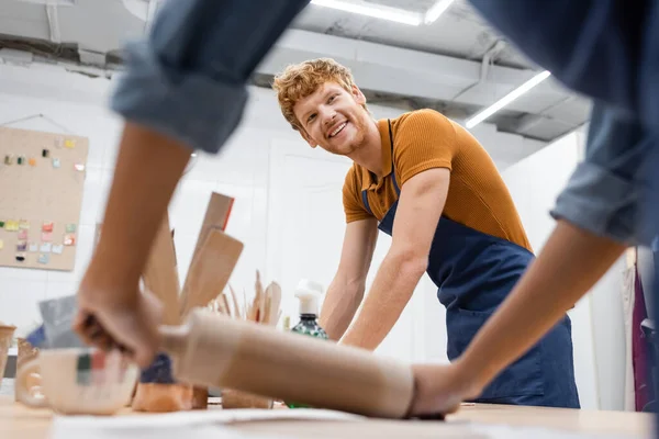 Homme rousse joyeux tablier regardant femme afro-américaine floue pâte à modeler avec rouleau à pâtisserie — Photo de stock