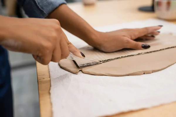 Vue recadrée d'une femme afro-américaine coupant de l'argile avec couteau près du carton — Photo de stock