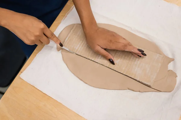 Vista superior de la mujer afroamericana cortando arcilla con cuchillo cerca de cartón - foto de stock