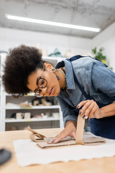 Femme afro-américaine bouclée dans des lunettes déchirant carton pendant le cours de poterie — Photo de stock