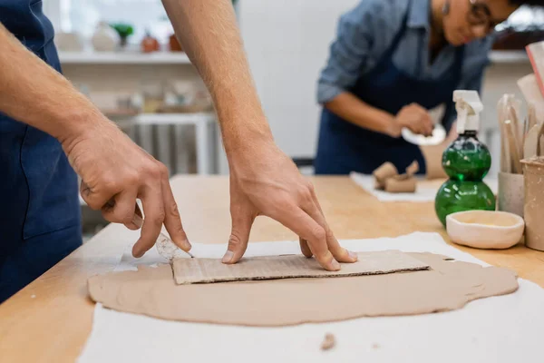 Abgeschnittene Ansicht eines Mannes, der in der Nähe einer Afroamerikanerin mit einem Messer Ton um Karton schneidet, auf verschwommenem Hintergrund — Stockfoto