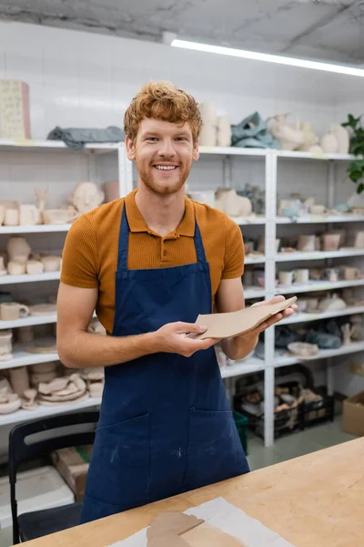 Cheerful redhead man with beard standing in apron and holding rectangle shape clay piece in hands — Stock Photo