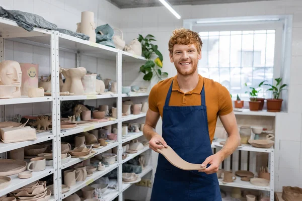 Positive redhead man in apron holding rectangle shape clay piece in hands near sculptures on rack — Stock Photo