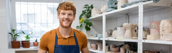 Pelirrojo positivo en delantal sonriendo cerca de esculturas en estantes de estante en la tienda de cerámica, pancarta - foto de stock