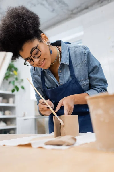 Glücklich afrikanisch-amerikanische Frau in Schürze hält Shaper während Modellierung Ton in Töpferei-Werkstatt — Stockfoto