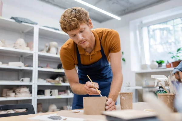 Bearded man in apron holding shaper while modeling clay during pottery class — Stock Photo