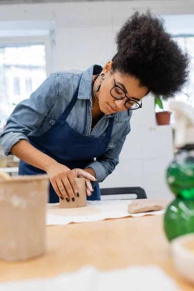 Молодая африканская американка в фартуке, моделирующая глиняные изделия во время урока керамики — стоковое фото