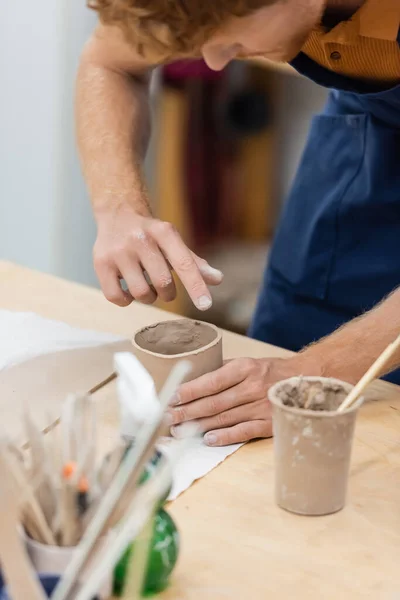 Focused man with red hair shaping clay piece into cup during pottery class — Stock Photo