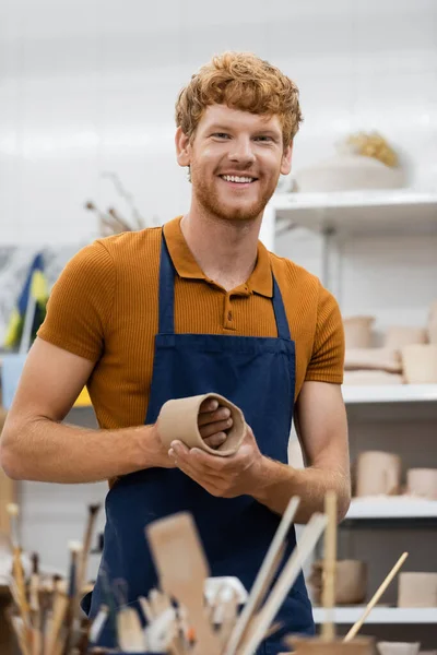 Happy and bearded man in apron shaping clay piece into cup during pottery class — Stock Photo