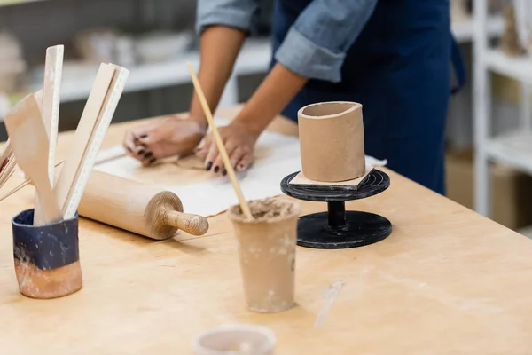 Clay cup on stand near rolling pin and african american woman handcrafting in studio — Stock Photo