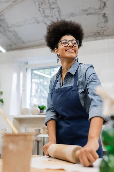 Cheerful african american woman in eyeglasses and apron modeling clay with rolling pin — Stock Photo