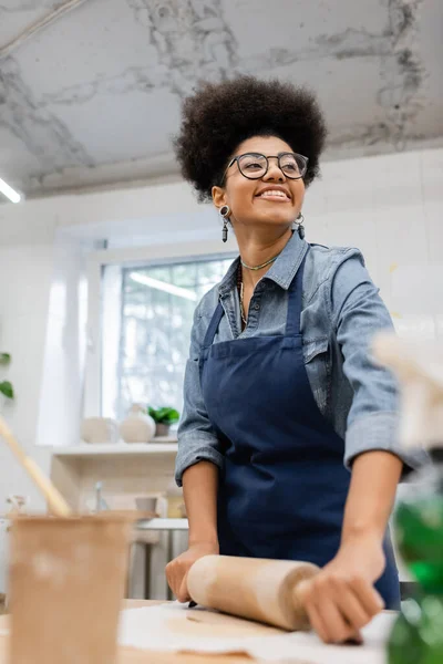 Alegre afroamericana mujer en gafas y delantal modelado de arcilla con rodillo - foto de stock