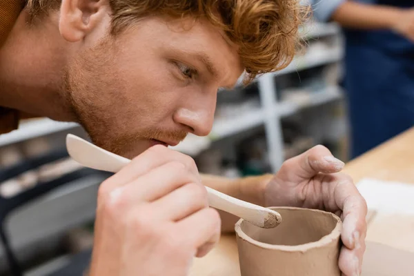 Concentrated man with red hair holding shaper while modeling clay cup in pottery workshop — Stock Photo
