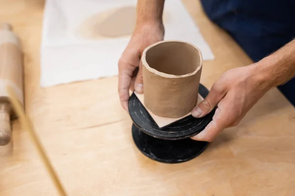 Vista ritagliata dell'uomo che tiene il basamento con la tazza di argilla nell'officina ceramica — Foto stock