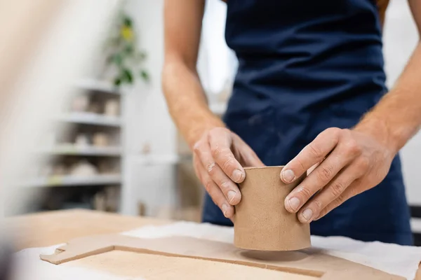 Vista ritagliata dell'uomo in blu grembiule stampaggio tazza di argilla in laboratorio di ceramica — Foto stock