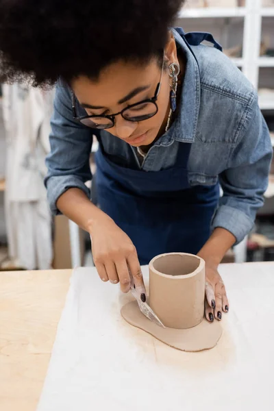 Curly african american woman in eyeglasses cutting clay around shaped cup with knife during pottery class — Stock Photo