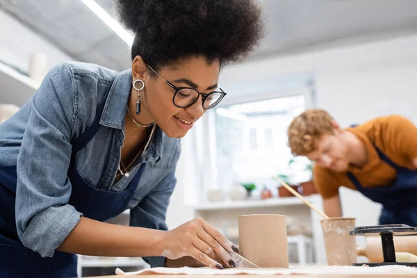 Glücklich afrikanisch-amerikanische Frau schneidet Ton um geformte Tasse mit Messer in der Nähe verschwommenen Mann während Töpferkurs — Stockfoto