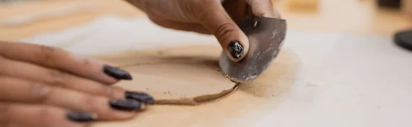 Vista recortada de la mujer afroamericana con manicura que sostiene rascador de acero inoxidable mientras moldea la pieza de arcilla, bandera - foto de stock