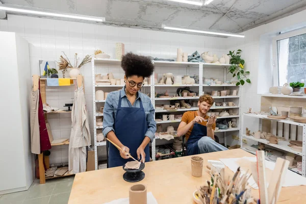 Cheerful and interracial man and woman in aprons handcrafting during pottery lesson — Stock Photo